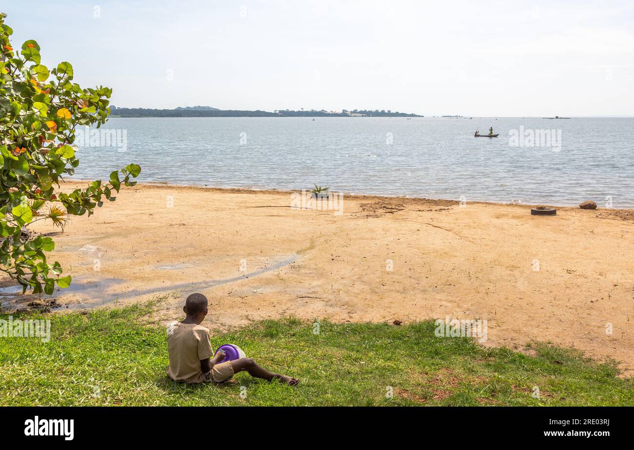 Young boy sitting on the edge of the beach watching a fishing boat in the distance. Sienna Beach, Lake Victorie, Entebbe. Stock Photo