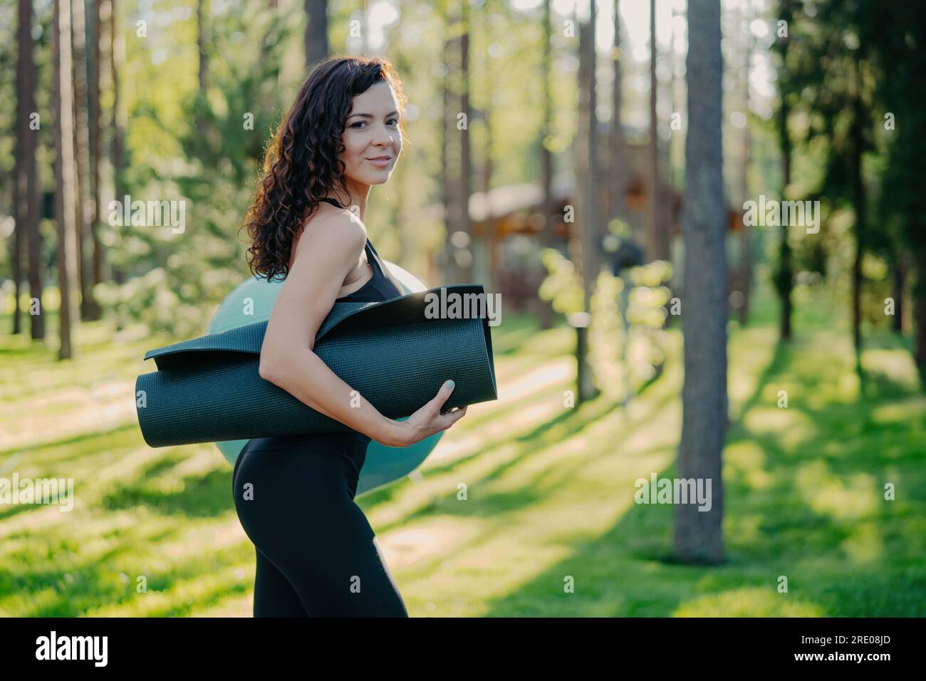Slim sporty woman carries karemat under arm, dressed in black leggings. Poses against green grass and trees, ready for fitness ball exercises. Stock Photo