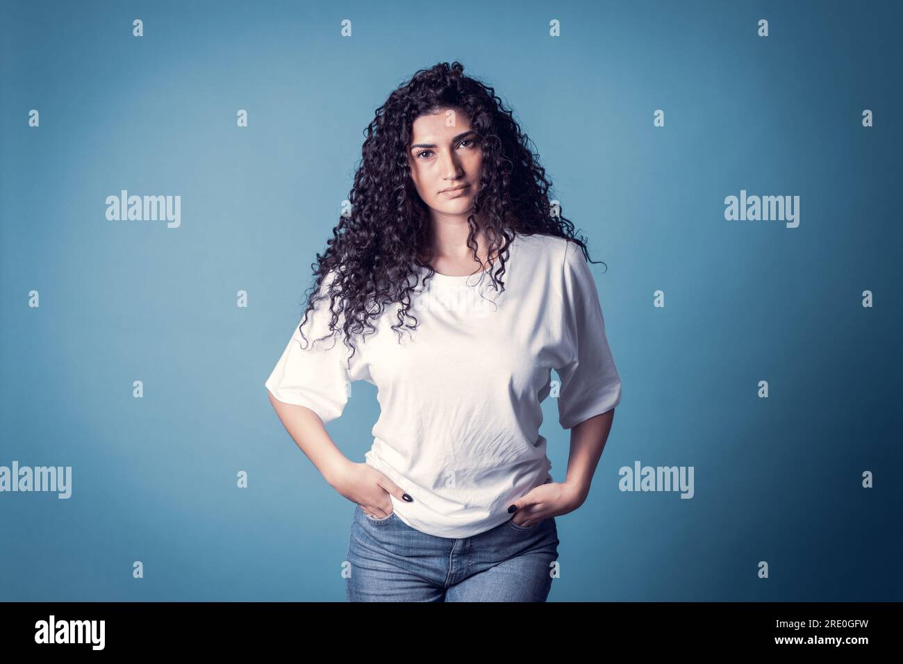 Shot of serious confident curly woman, wears proudly casual basic solid white t-shirt isolated over blue background. Stock Photo