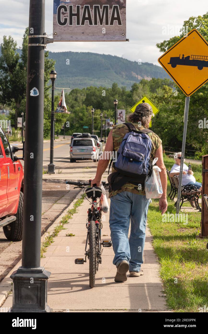 A man in casual dress and a backpack walks his bike down the sidewalk in downtown Chama, New Mexico, United States. Stock Photo