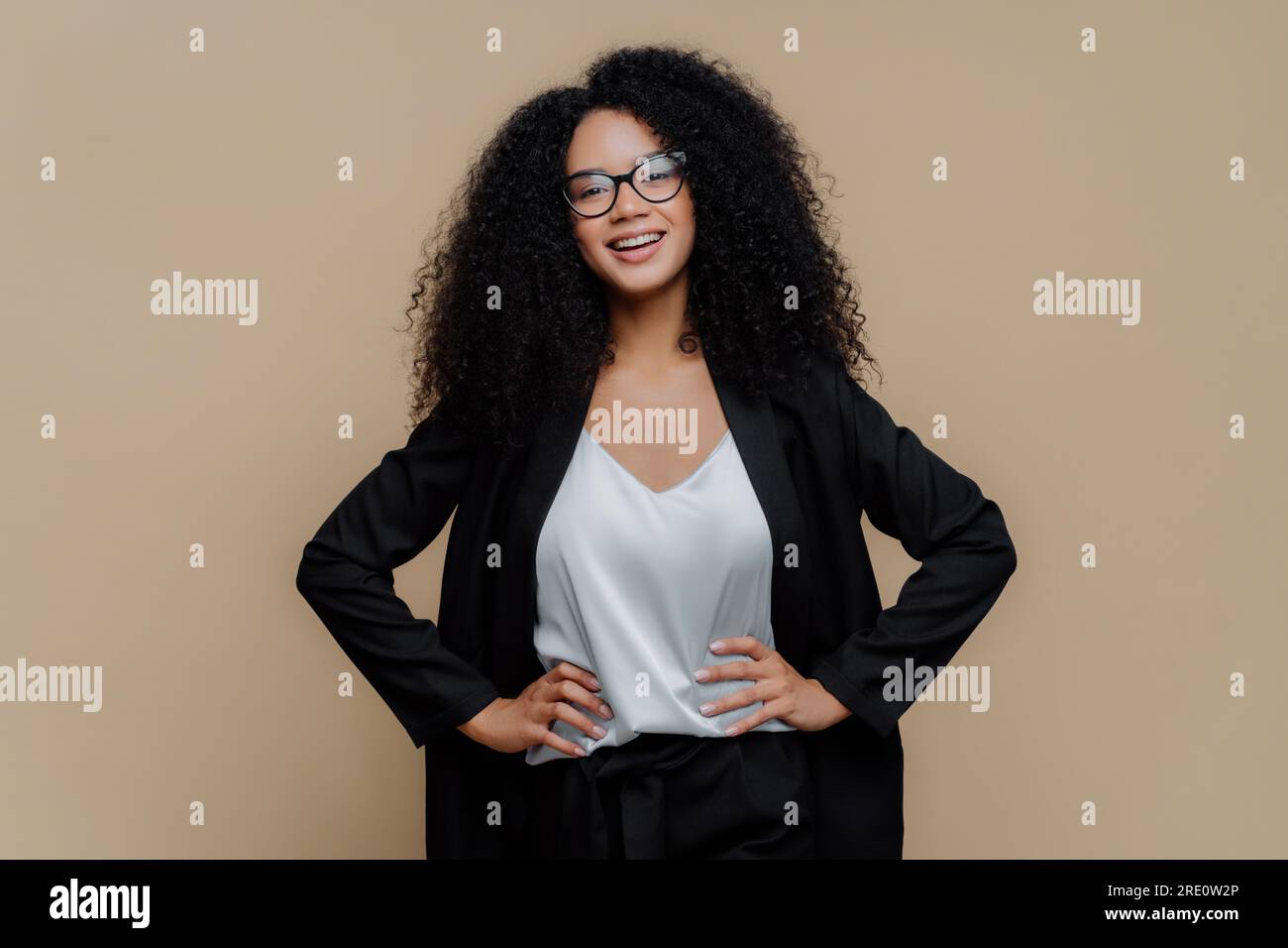 Joyful Afro woman, elegant black clothes, hands on hips, glasses, beaming smile. Female director, isolated on brown. Stock Photo