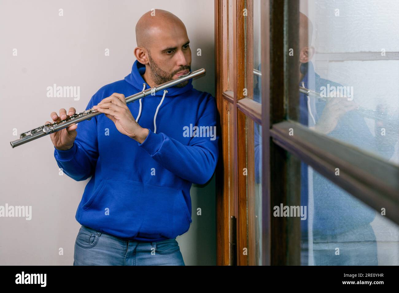 young latino young man in blue, inspired and melancholic playing the flute at home and standing and looking out the window. Stock Photo