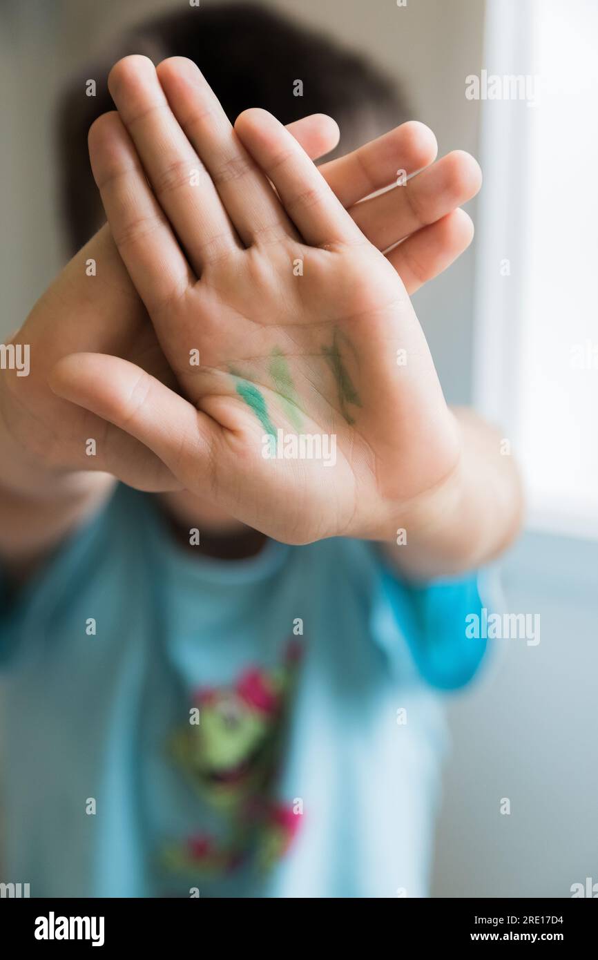 Boy hiding his face and showing his hands after doing painting activities. Concept of learning, skills, happy childhood. Focus on hands. Stock Photo