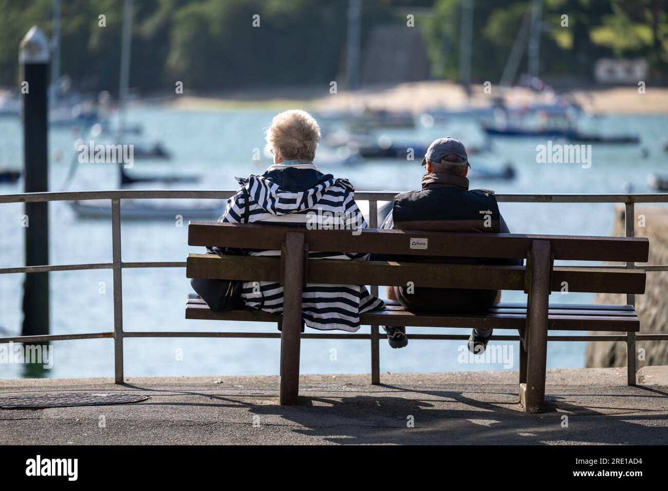 Couple sitting on a public bench in Whitestrand car park, in the centre of Salcombe, Devon, overlooking moored boats in the harbour on a sunny day Stock Photo