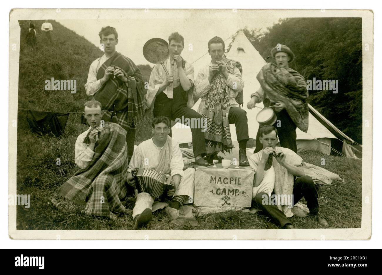 Original early 1920's era postcard of group of good looking young men, messing about whilst camping dressed as Scottish pipers and bandsmen. One of them holds up a greasy frying pan with a charred sausage stuck to it. The lads are wearing tartan - one has a sporran made from a  hair brush attached to the front of his kilt! They have a sign outside their tent stating 'Magpie camp' , unknown location in U.K. possibly Scotland. Stock Photo