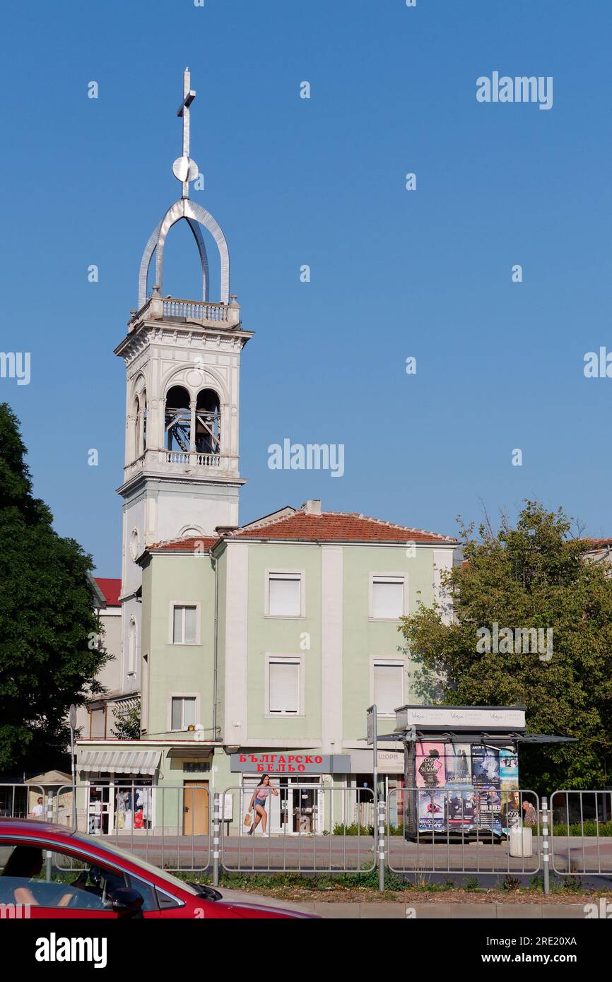Lady in pink top and short jeans walks in front of a Clothing shop rendered in green with church tower behind in Plovdiv, Bulgaria July 24, 2023 Stock Photo