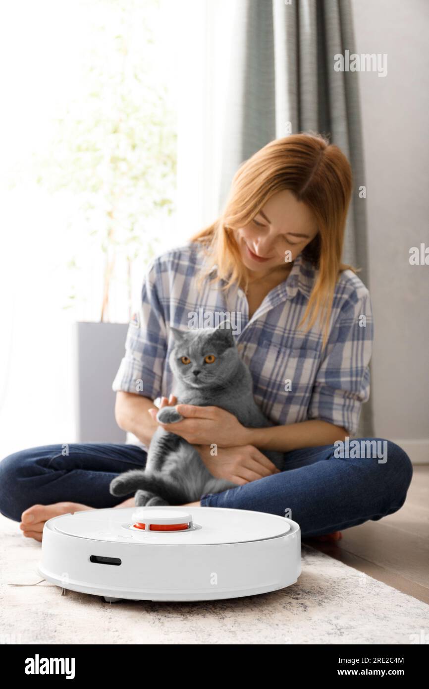 A smart robot vacuum cleaner cleans the carpet, a girl and a cat look at the vacuum cleaner. The concept of smart home, daily cleaning, allergy contro Stock Photo