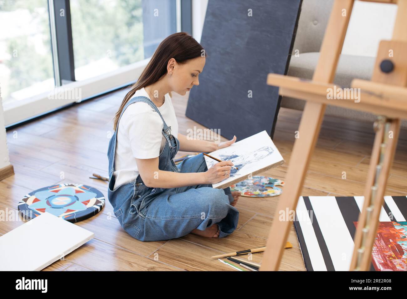 Side view of focused female painter wearing denim jumpsuit sitting on  wooden floor and working on new drawing at own art studio. Young lady  spending free time with canvas and paintbrush in