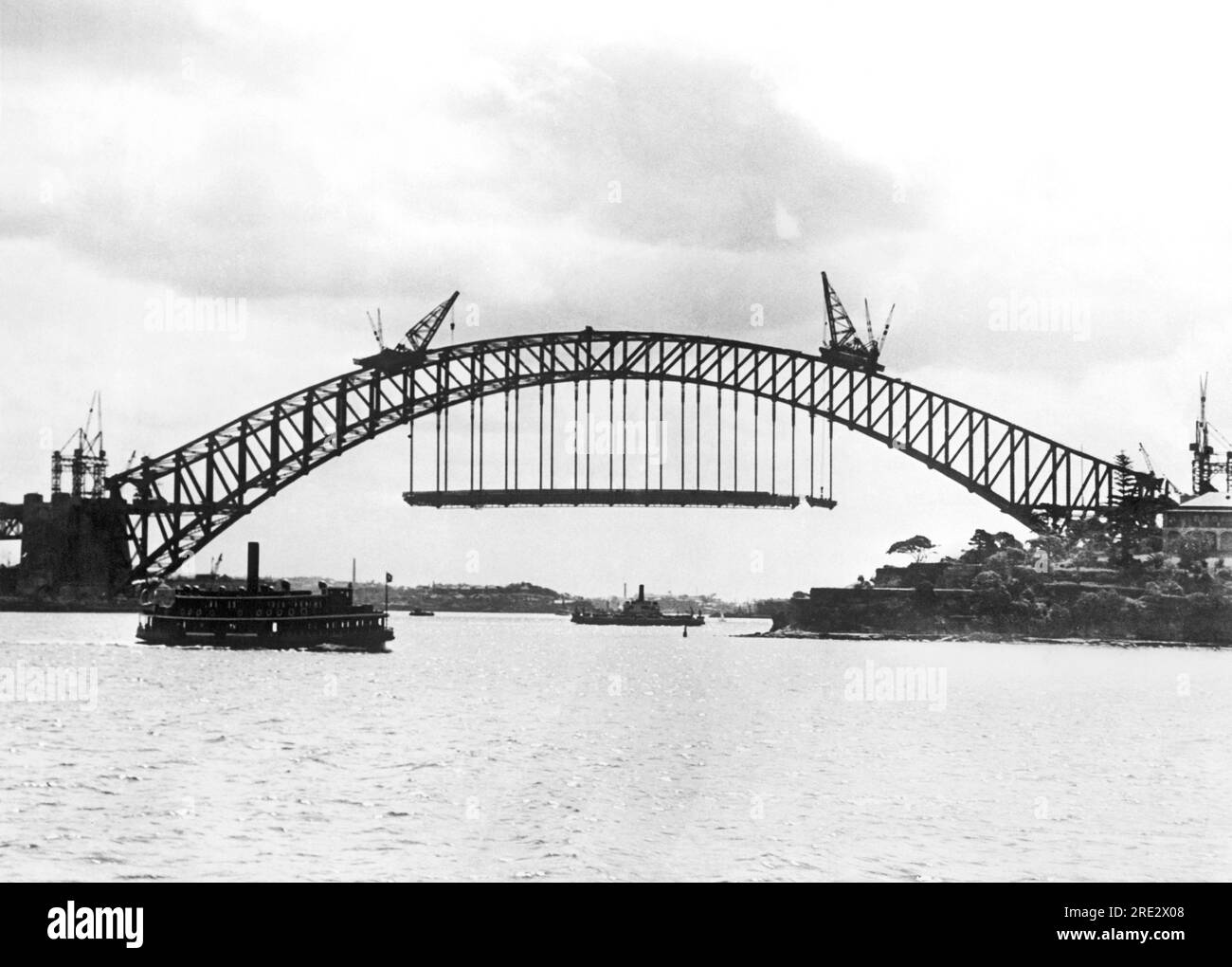 Sydney, Australia:  c. 1930 A vehicle ferry passes by the Sydney Harbour Bridge under construction between the Sydney business district and the North Shore. Stock Photo