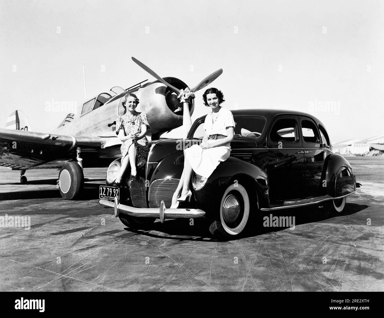 California:  1939 Two women sit on the fenders of a 1939 Lincoln Zephyr sedan at an airport with a single engine plane parked behind them Stock Photo
