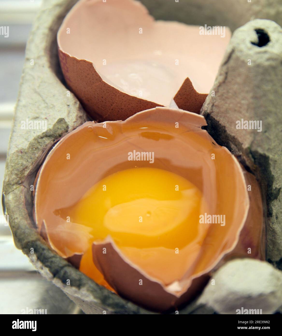 Yolk and white of a broken egg in its half shell with rest of shell next to it, sat in a cardboard eggs carton, close up Stock Photo