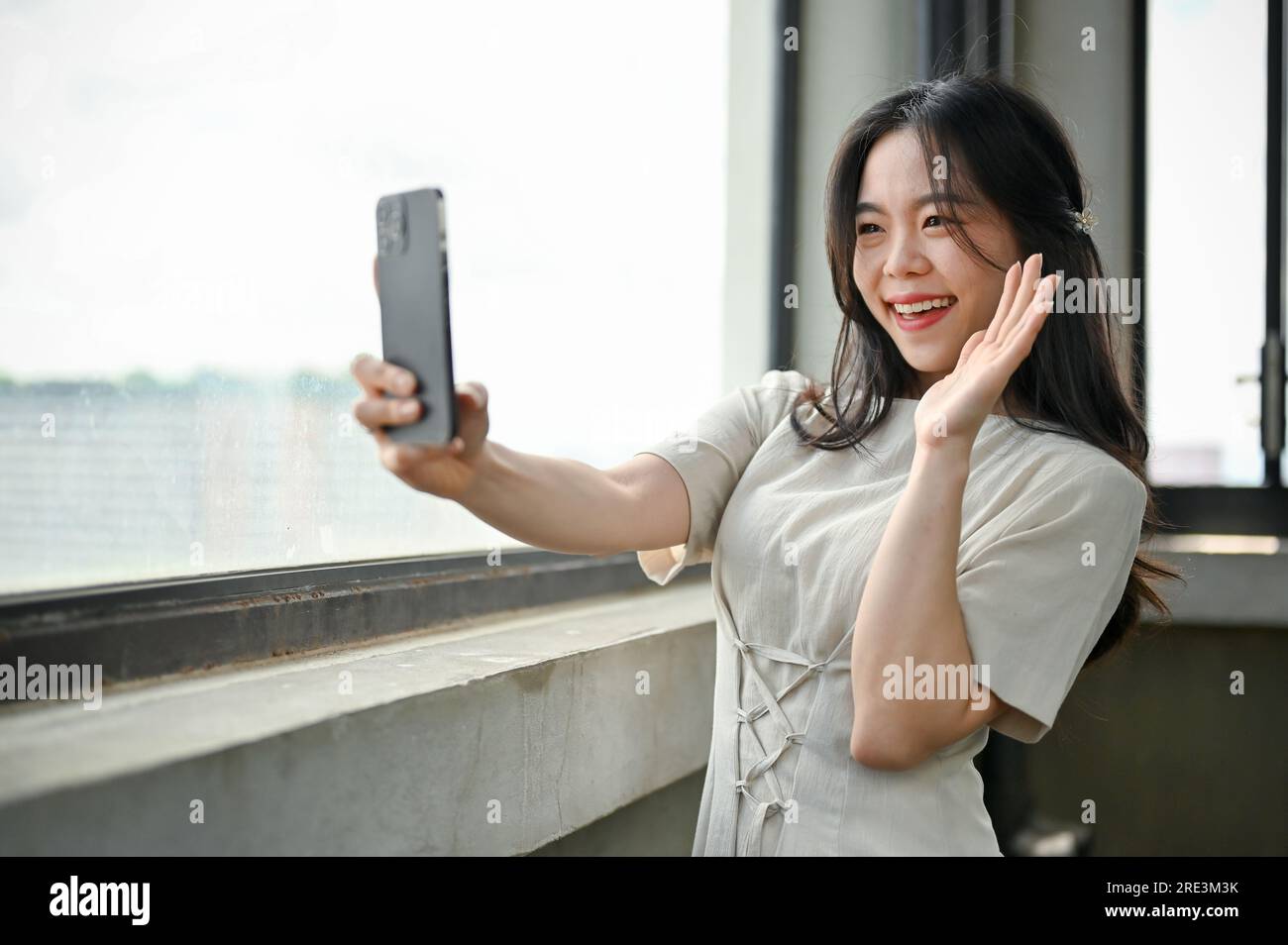 An attractive Asian woman in a beautiful dress is taking selfies with her smartphone while standing by the window in a building. Stock Photo