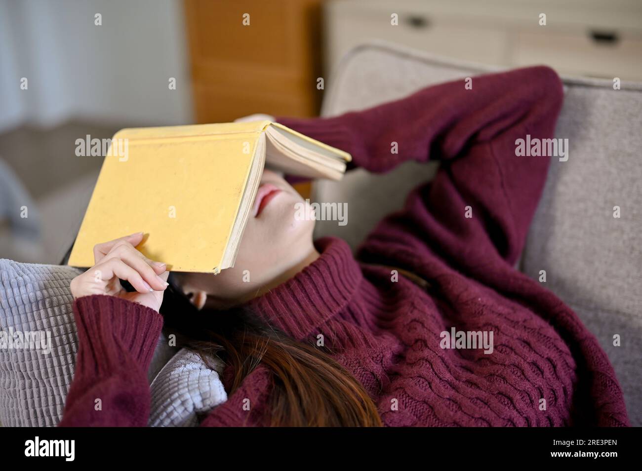 Young Asian woman laying down on the sofa with book cover on her face, taking a nap, relaxing in free time Stock Photo