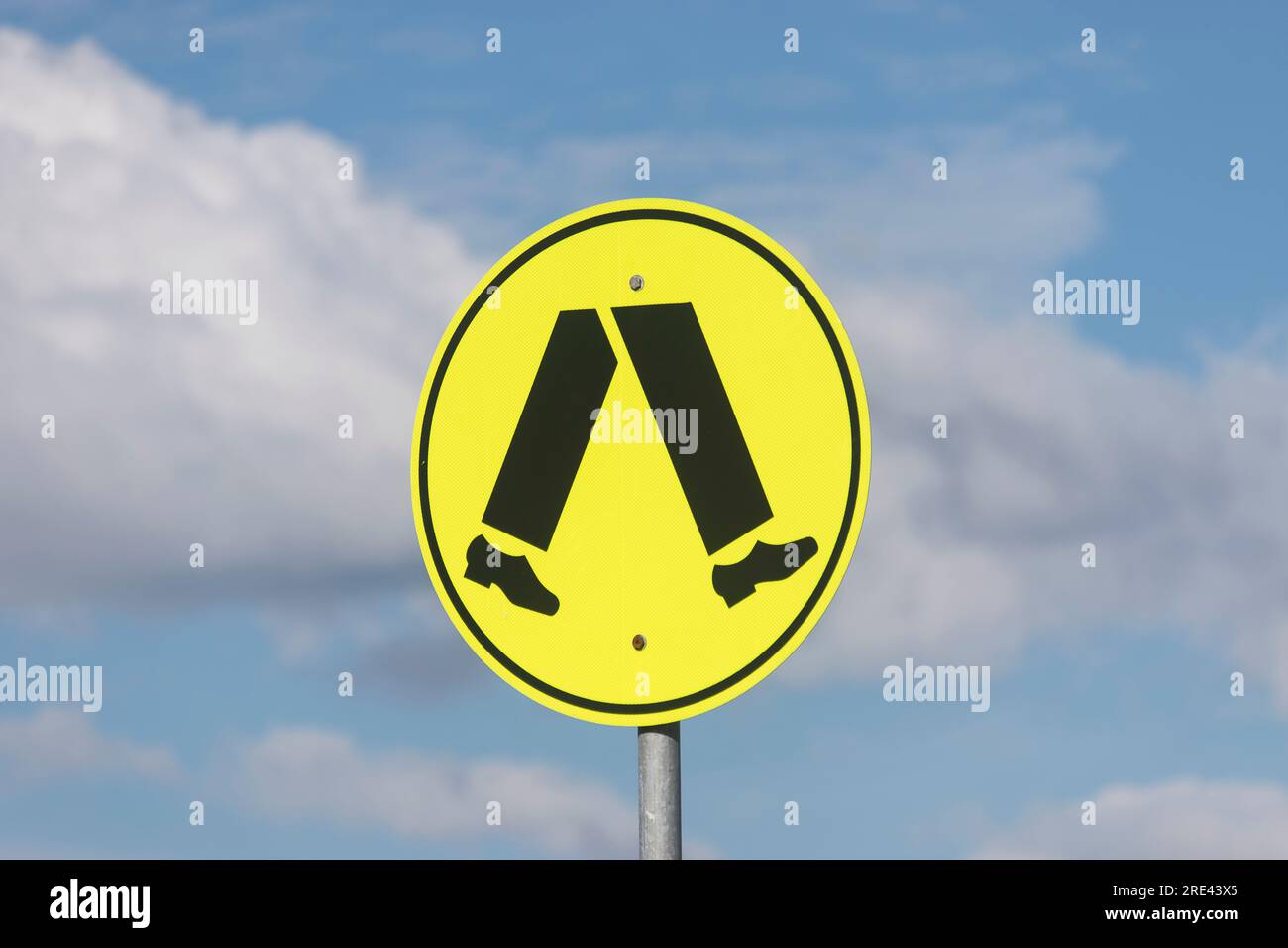Black and luminous yellow circular road sign at pedestrian crossing. Legs and feet walking. Blue sky and white clouds above. Queensland, Australia. Stock Photo