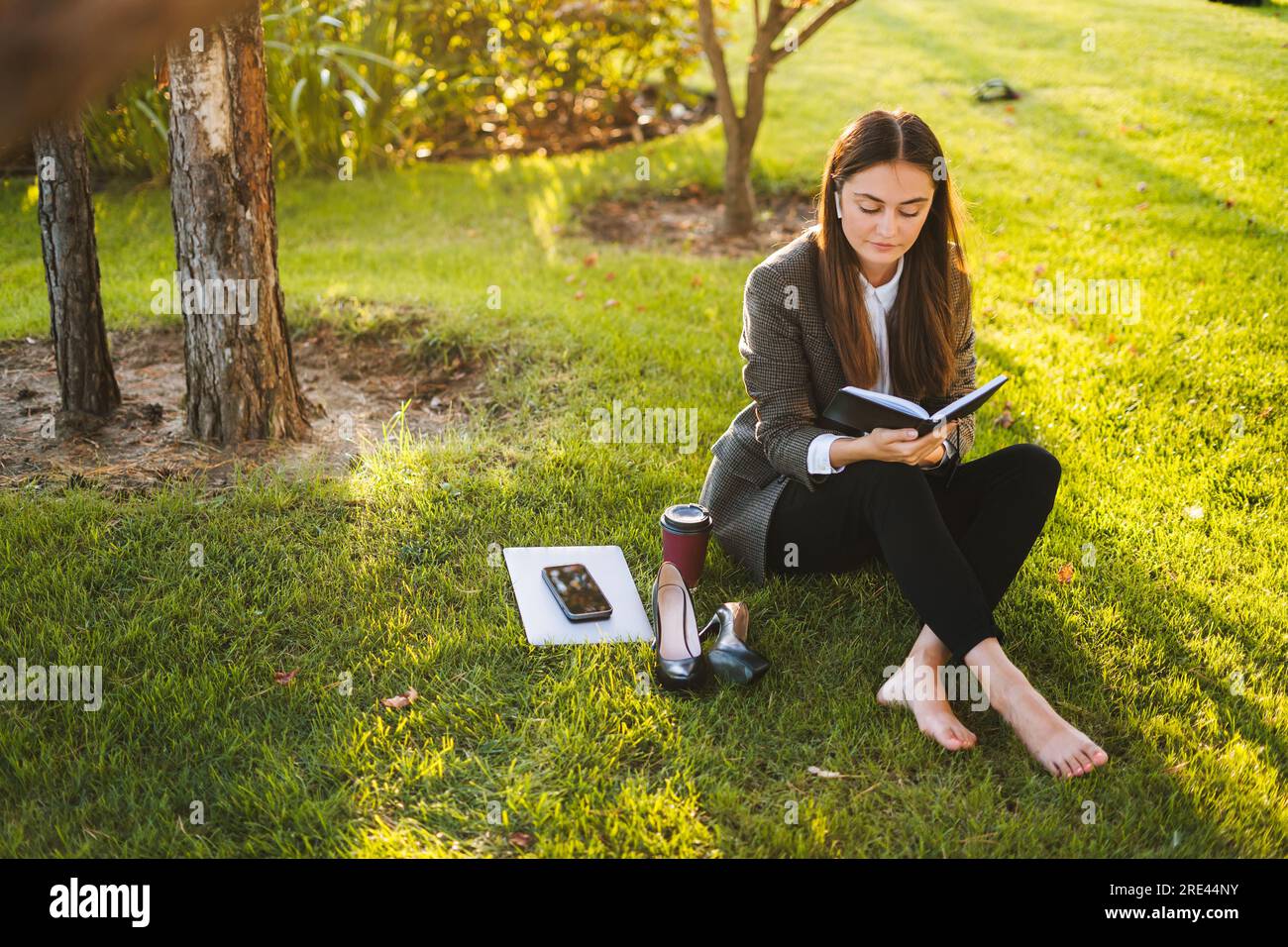 Young beautiful calm woman in a jacket reading a book in a summer green downtown. Urban lifestyle. Young business woman. Stock Photo