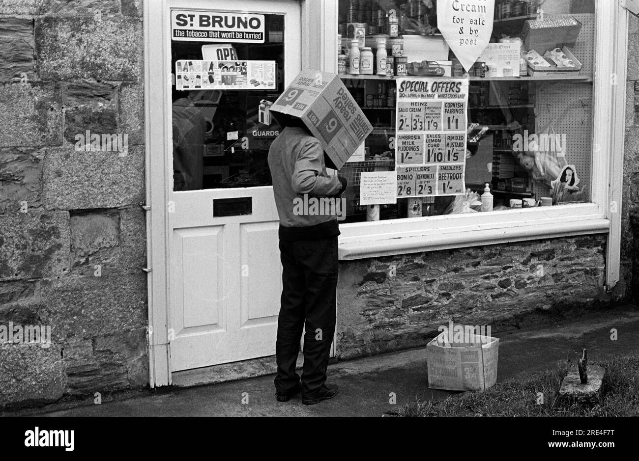 Village shop 1970s UK. A young boy with empty cardboard box of Ready Salted Crisps over his head outside a village store, grocery shop, which is selling 'Cream for sale and by post.' 'St Bruno the tobacco that wont be hurried.' Guards cigarettes. The shops special offers include, ' Heinz Salad Cream, Cornish Fairings, Ambrosia Milk Puddings, Gales Lemon Curd, RMixed Mustard, Spillers Bonio and Kenomeat and Smedleys Beetroot.'  Children's plastic dolls come in a clear plastic bag and hang in the window. They also sell a variety of ice creams. St Just, Cornwall. 1970 England. HOMER SYKES. Stock Photo