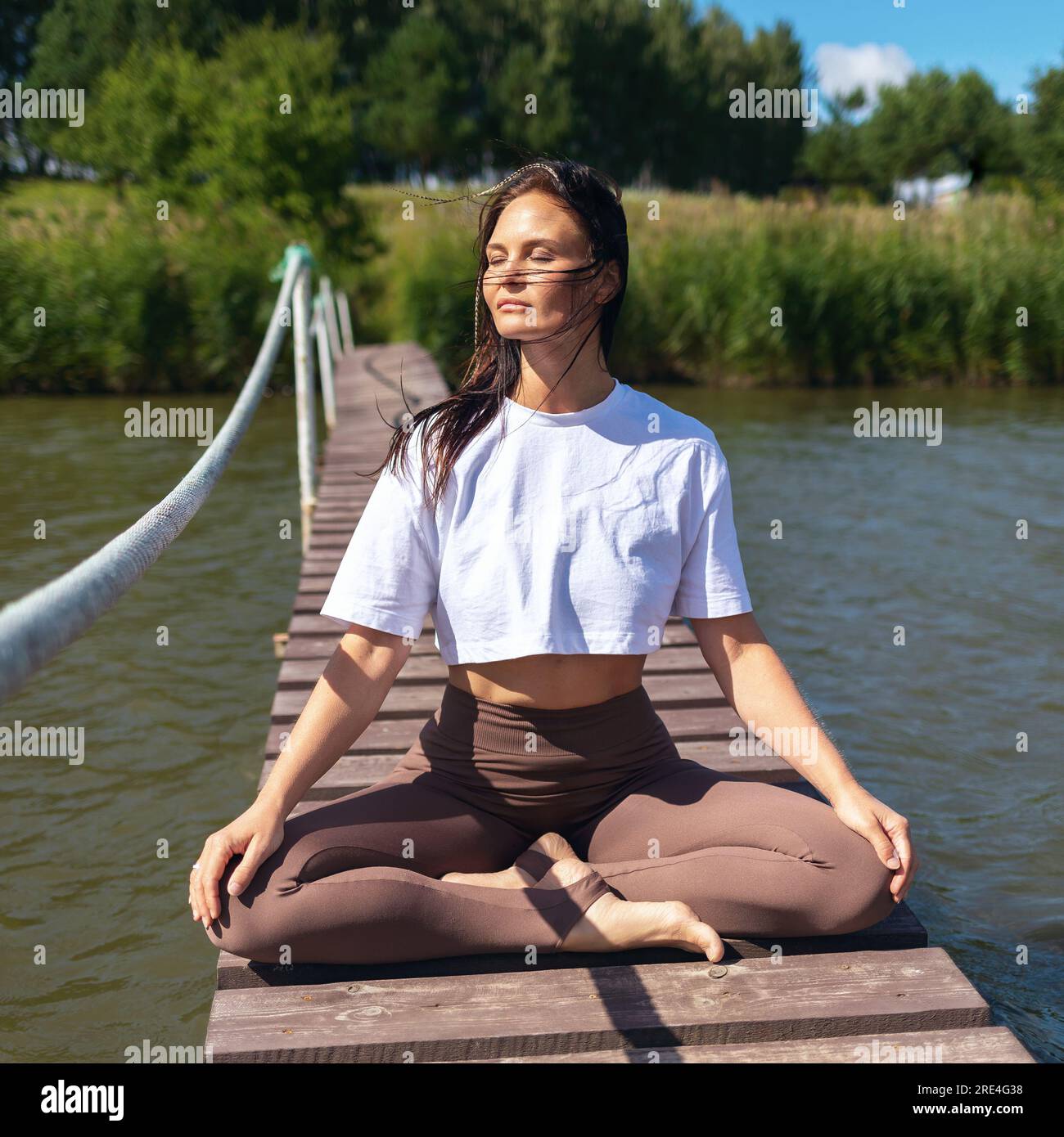 A woman practicing yoga, meditating sitting in a lotus position on a wooden bridge near the lake, training in sportswear Stock Photo