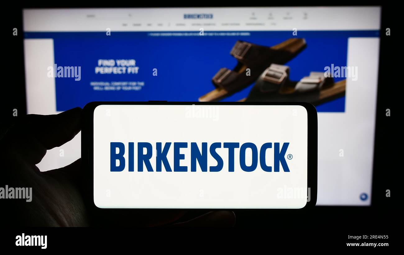 Person holding mobile phone with logo of shoe company Birkenstock Group B.V. Co. KG on screen in front of web page. Focus on phone display. Stock Photo