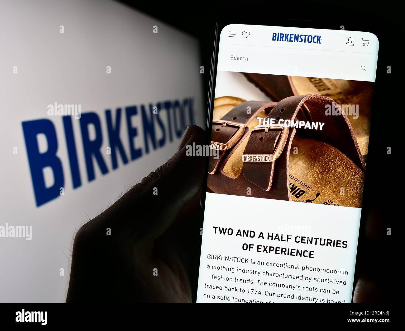 Person holding mobile phone with website of shoe company Birkenstock Group B.V. Co. KG on screen with logo. Focus on center of phone display. Stock Photo