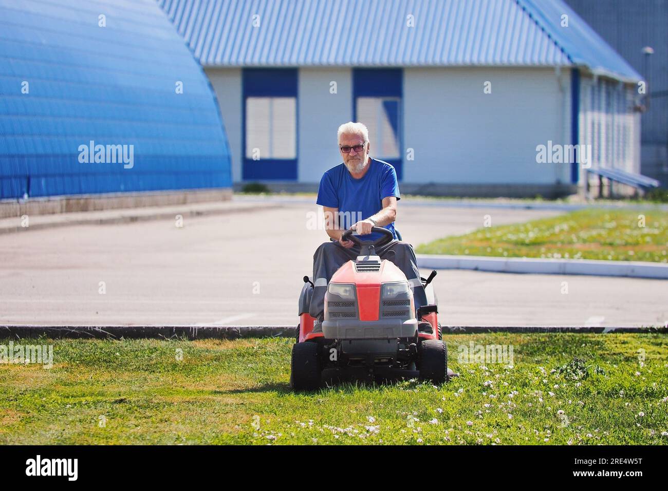 Elderly handyman gardener mows lawn on lawn mower tractor. Elderly man works as laborer. Mowing territory of industrial facility on summer day. Real workflow. Stock Photo