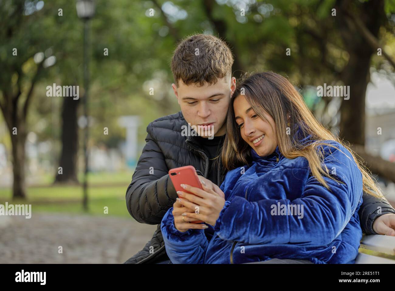 Young couple looking at a mobile phone sitting on a bench in a public park. Stock Photo