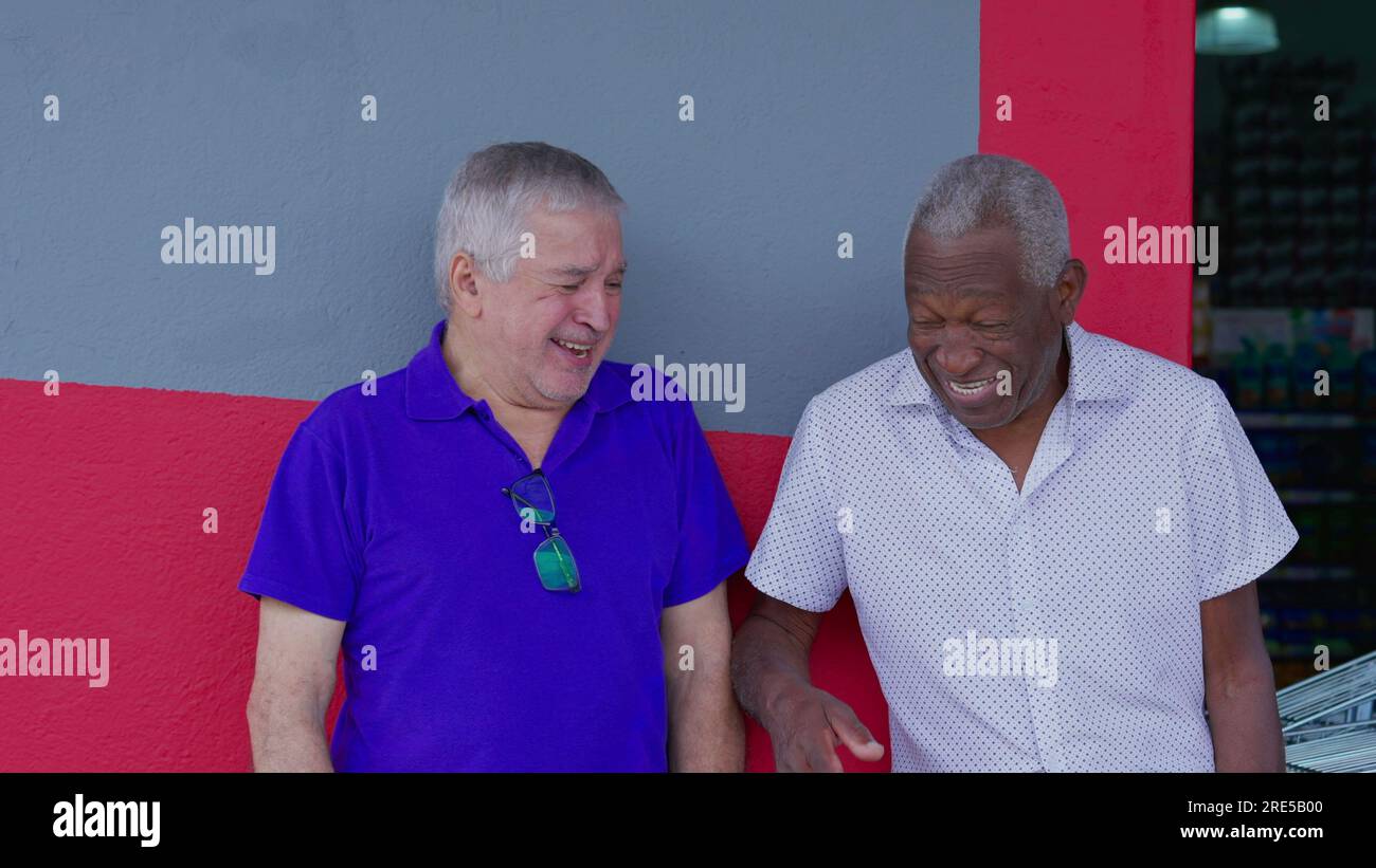 Joyful two diverse male senior friends smiling and laughing together leaning on sidewalk wall. A caucasian and African American older men interaction Stock Photo