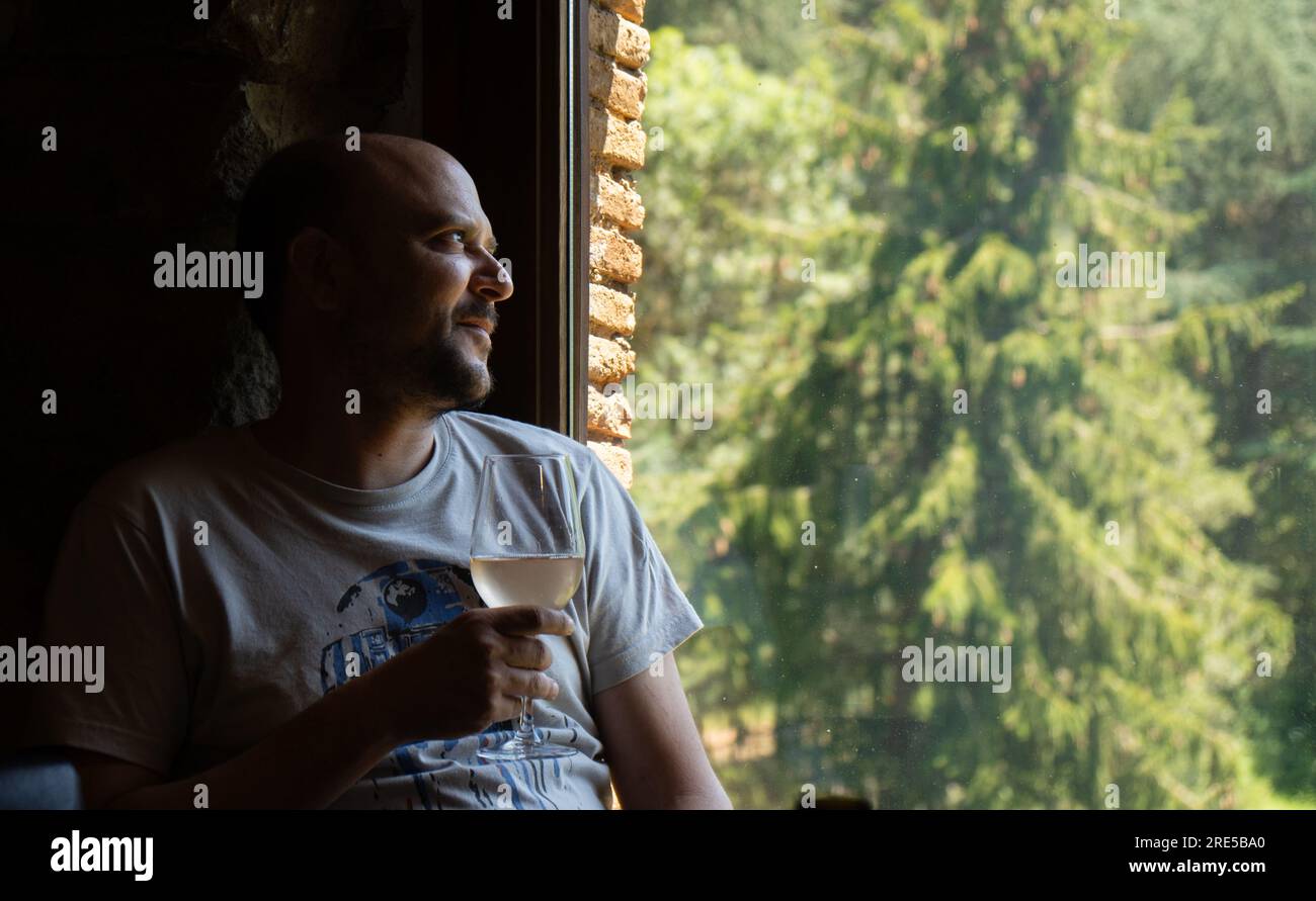 Relaxed middle-aged man with a glass of white wine in his hand illuminated by a dim evening light looking out a window of a rural house with green tre Stock Photo