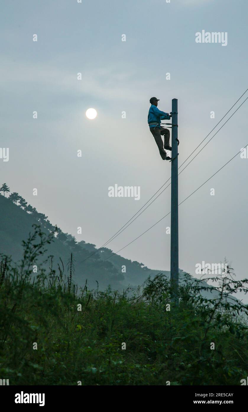 A silhouette of a worker climbing on a power pole to install hanging wires while the sun is barely visible through the smog and clouds, China Stock Photo