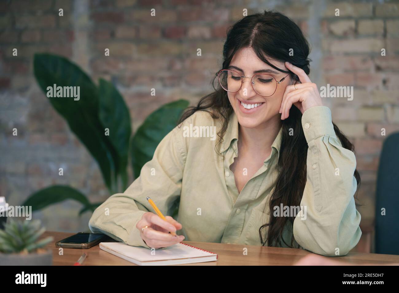 Smiling young brunette lady in glasses making notes in a notebook while working in the office. She working or studying. The startup, coworking Stock Photo