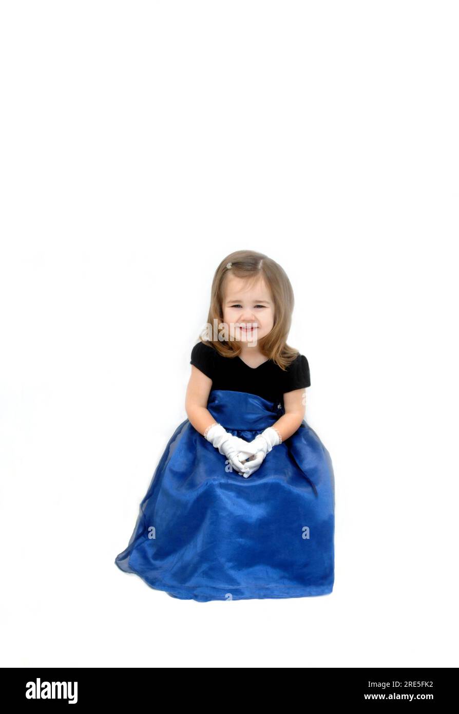 Little girl is wearing a royal blue gauze dress with velvet bodice.  She has long hair and is seated in an all white room. Stock Photo