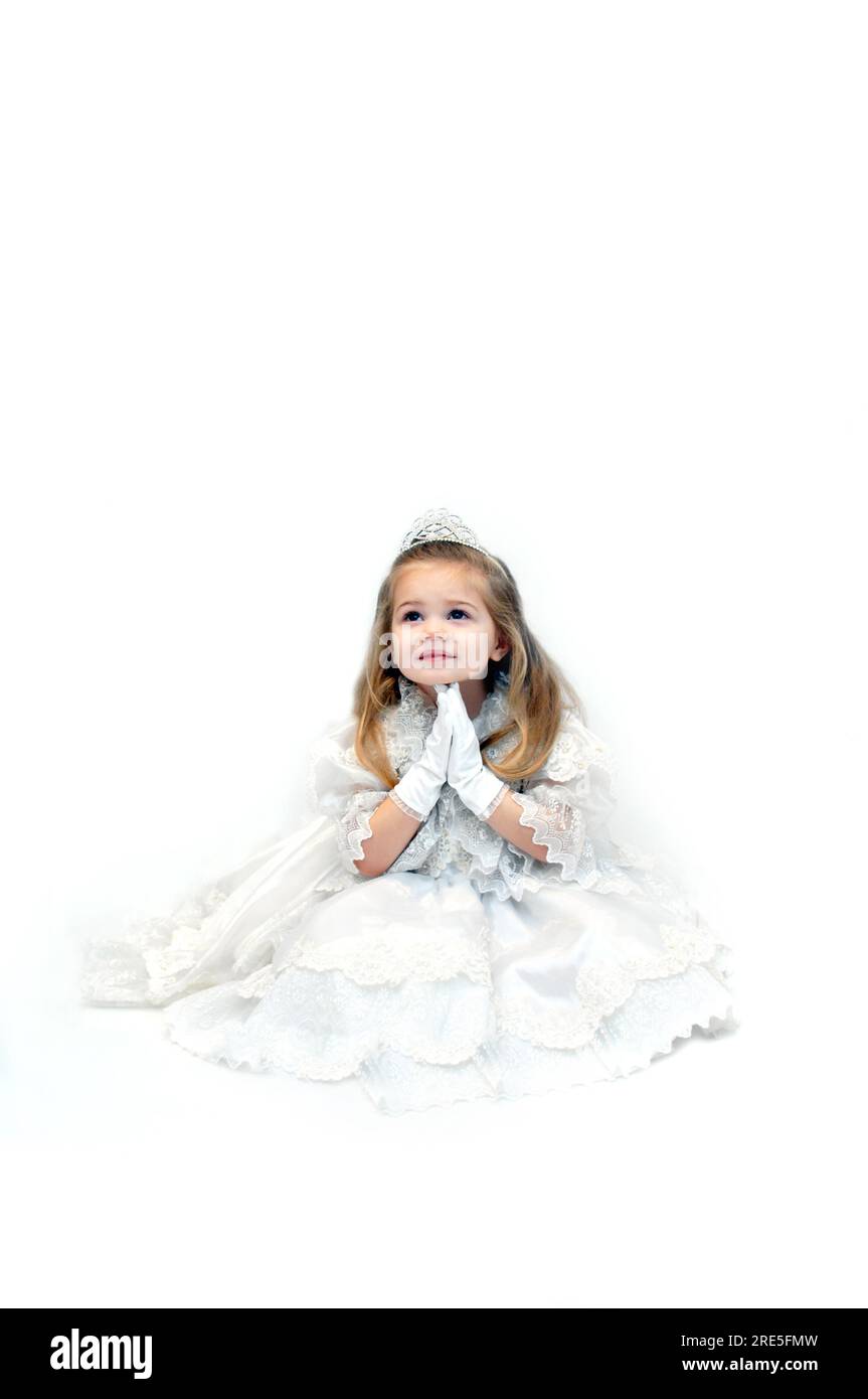 Little girl folds her hands in prayer.  She is sitting in an all white room and wearing an all white pageant dress and crown. Stock Photo