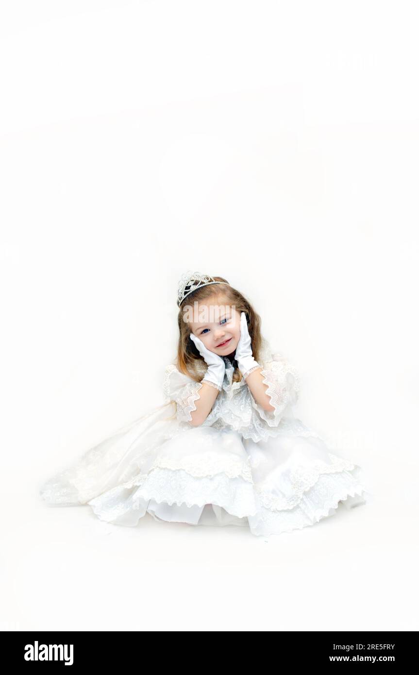 A little girl princes, dressed in elegant white gown and robe, wears a sparkling crown and holds her face in her gloved hands. Stock Photo