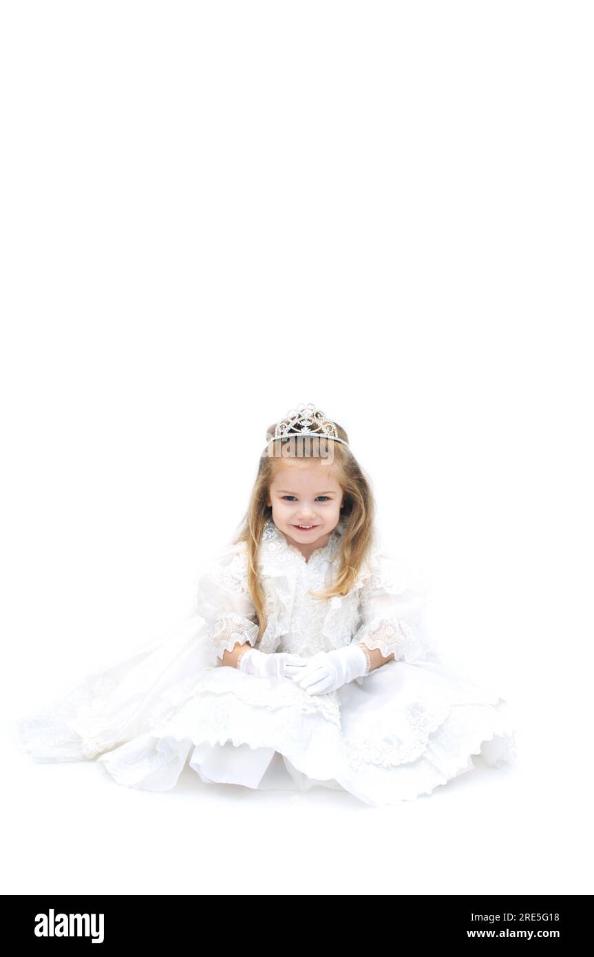 Wishful little girl is dressed up as a bride with sparkling crown tiara.  She is smiling happily and sitting in an all white room. Stock Photo