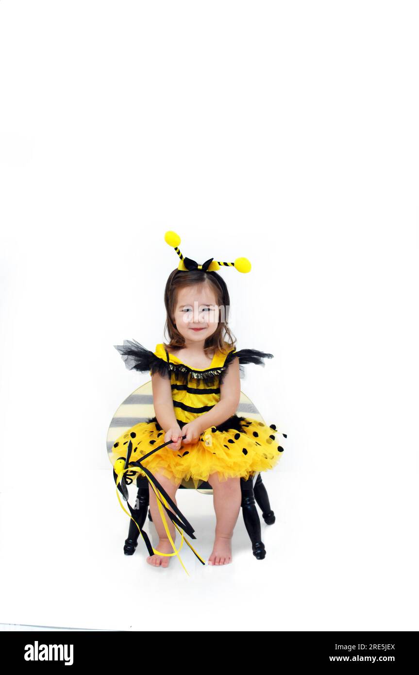 Little bumble bee sits on a black stool in an all white room.  She is holding a bumble bee wand and grinning. Stock Photo