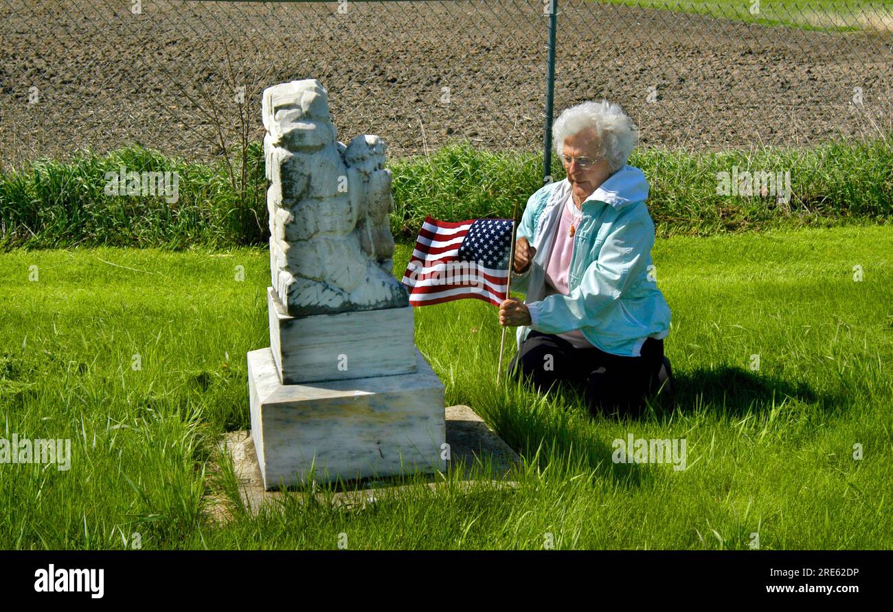 Mother places an American flag at the tomb of her loved one.  She is kneeling besides the grave on the grass. Stock Photo