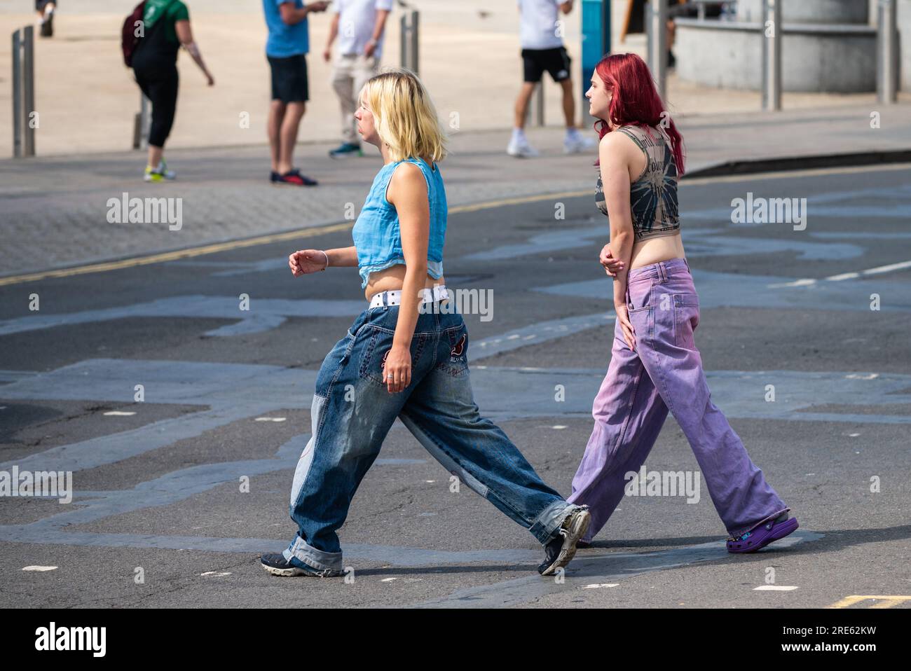 Pair of young slim women, girls or teenagers, dressed in jeans, walking across a road and going shopping in Summer, UK. Friends at shops. Stock Photo