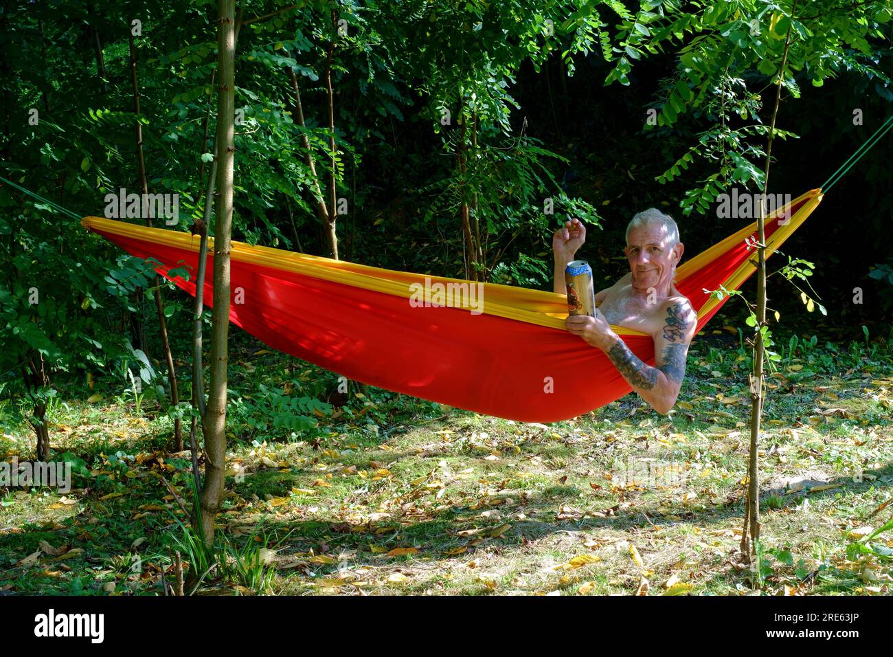 older man relaxing in garden hammock shaded by trees with cigarette and can of beer zala county hungary Stock Photo
