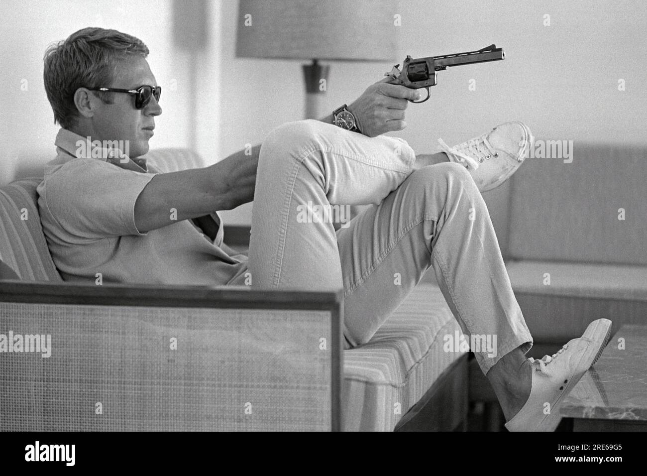 Profile view of American actor Steve McQueen (1930 - 1980) sitting on a sofa in his home in Palm Springs, California, sunglasses over his eyes as he aims a handgun. Steve McQueen, widely considered one of the coolest and most charismatic actors of the 1960s and 1970s, wears a white shirt and blue jeans. Steve McQueen looks focused and determined, with a slight twinkle in his eye and a firm grip around his gun. The background is indistinct and out of focus, creating a sense of isolation for the sitter. The overall mood of the image is one of toughness, confidence, and a sense of danger. Stock Photo