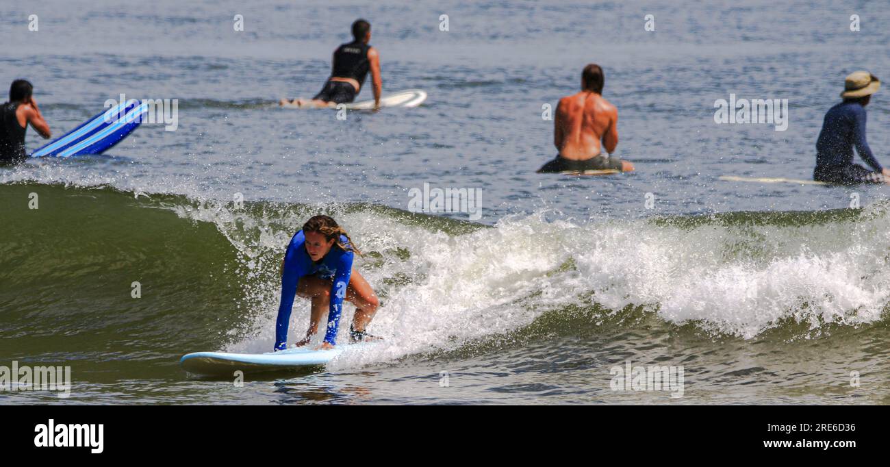 Gilgo Beach, New York, USA - 22 July 2023: Women surfing in a blue long sleeved rash guard riding a wave while others sit on their surfboards waiting Stock Photo