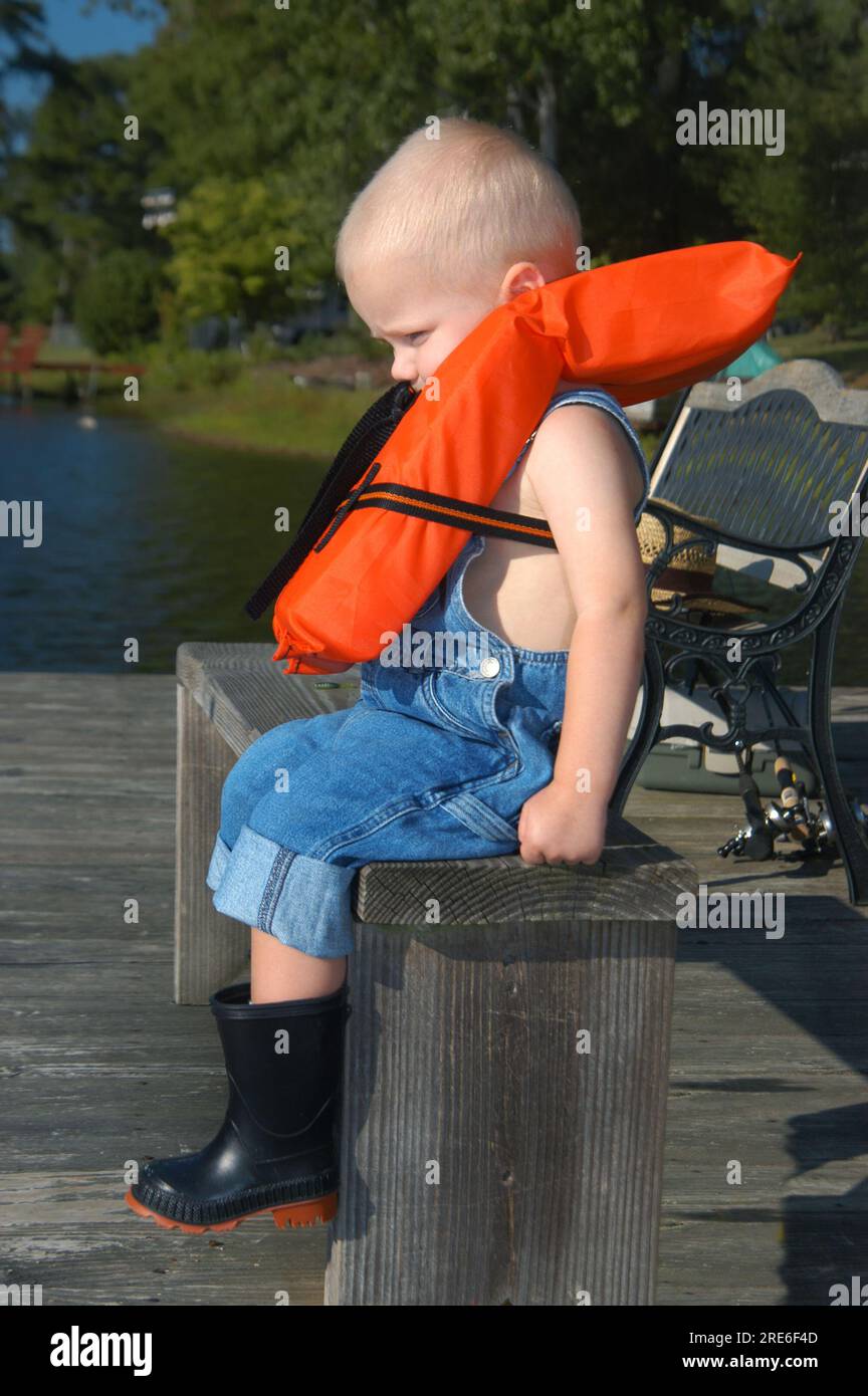 Little boy wears an orange, life-preserver as he sits on a rustic wooden bench on a fishing dock in Alabama.  He has on rolled up overalls and black r Stock Photo