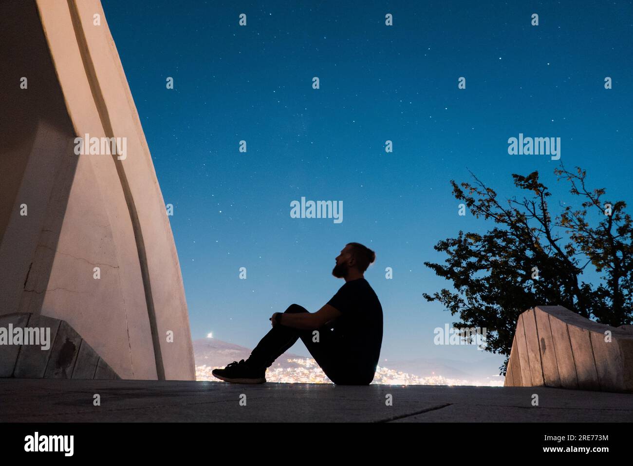 a man sits and looks at the night sky on the steps of the Ossuary Monument (Spomenik Kosturnica) in Veles, North Macedonia Stock Photo