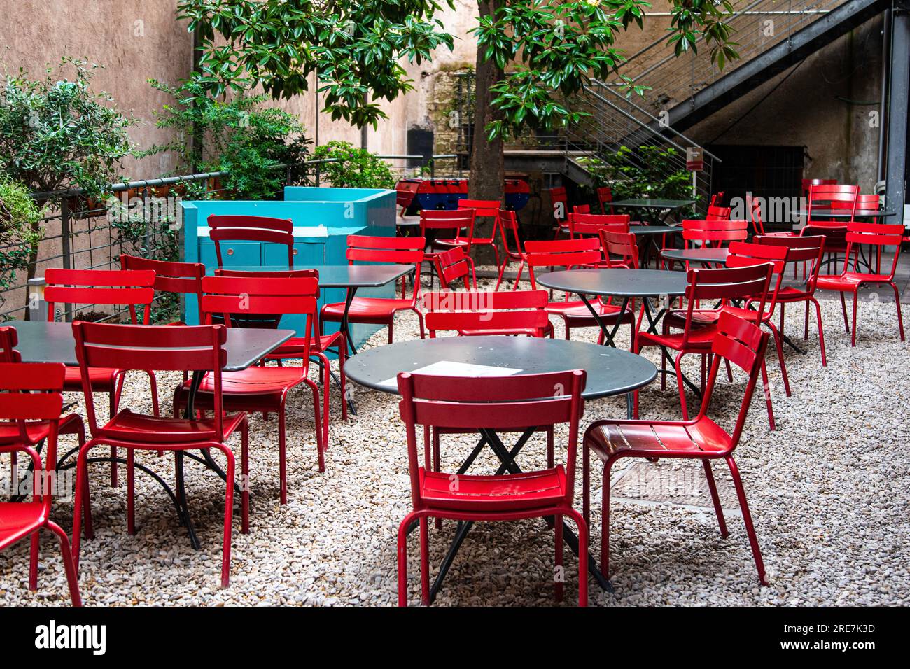 Bright red metal chairs around grey tables on a stone patio with trees
