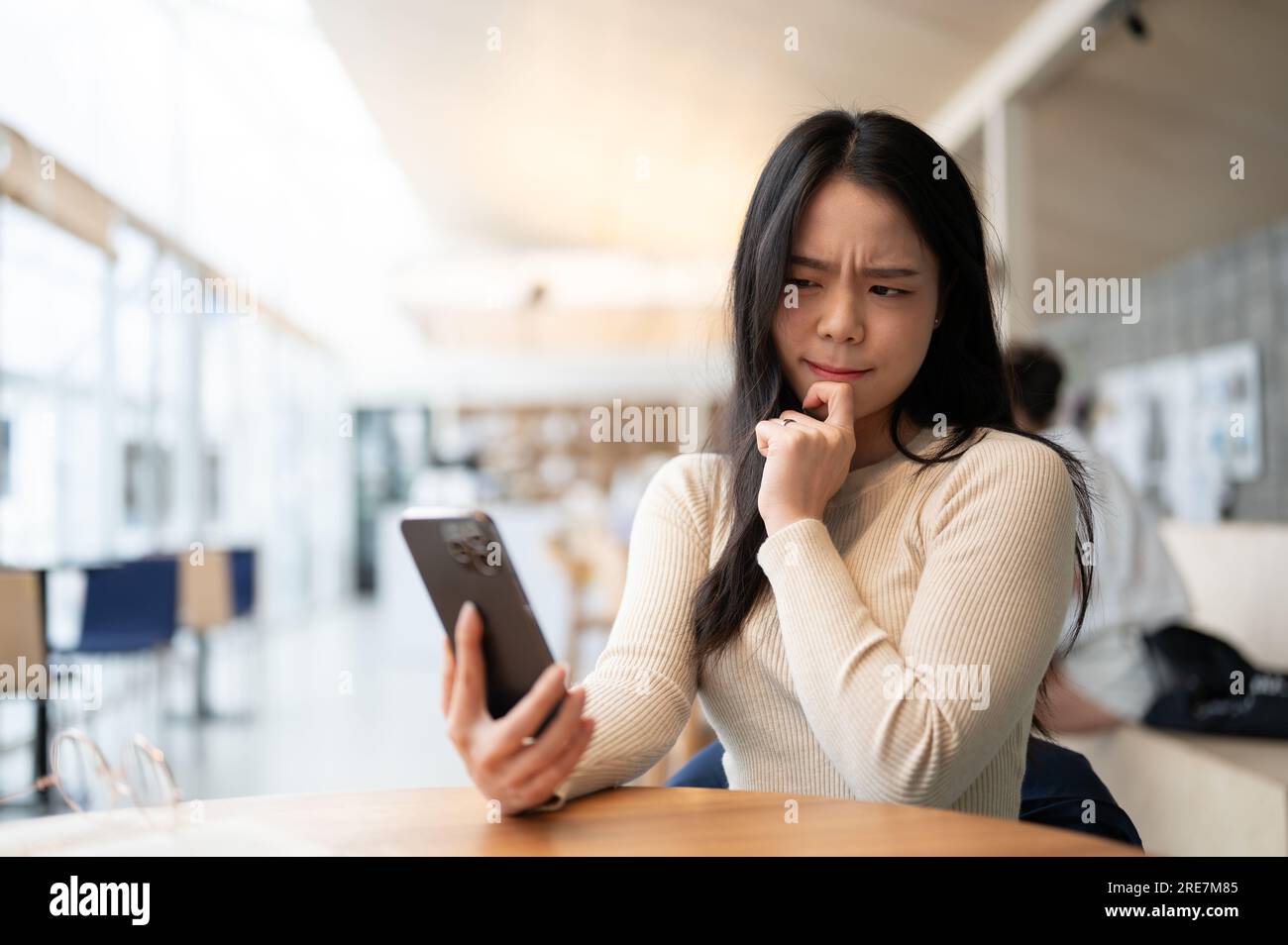 An unsure and thoughtful young Asian woman is looking at her smartphone screen with a serious and doubtful face while sitting in a coffee shop. worrie Stock Photo