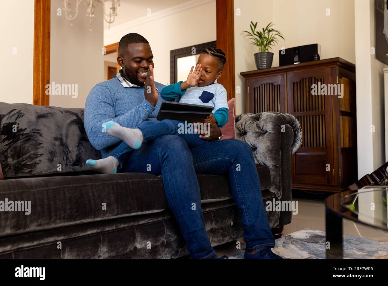 Happy african american father and son sitting in living room at home using tablet and high fiving Stock Photo