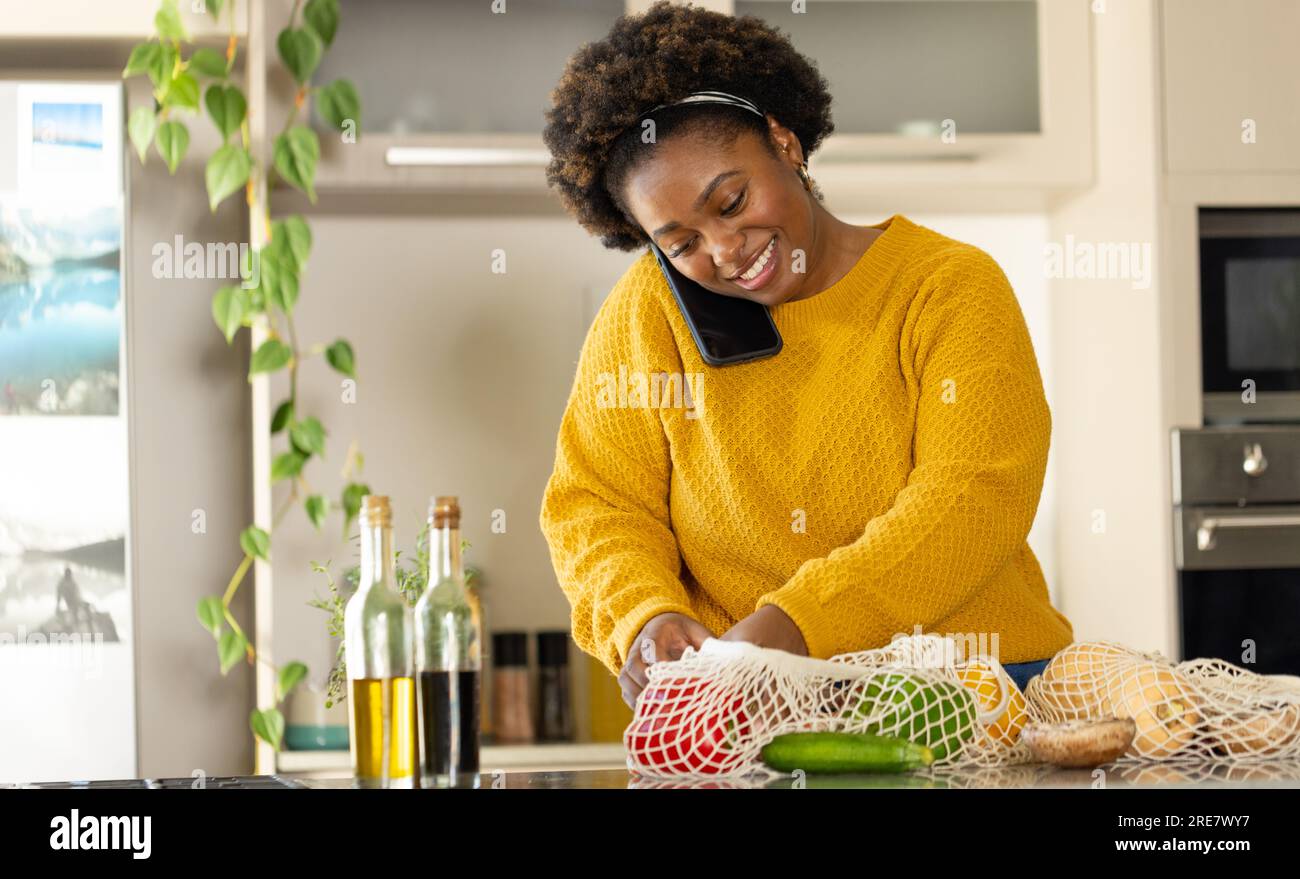 Happy plus size african american woman unpacking groceries and talking on smartphone in kitchen Stock Photo
