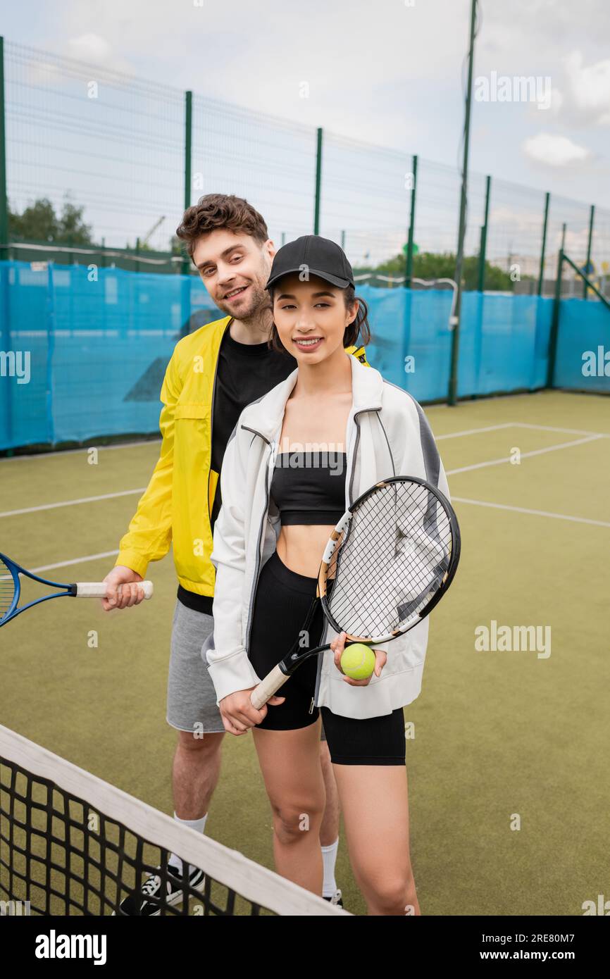 cheerful man and woman in active wear holding tennis rackets and ball ...