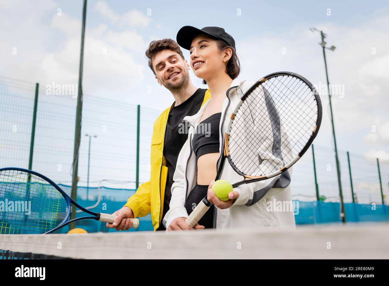 cheerful couple in stylish active wear holding tennis rackets and ball ...