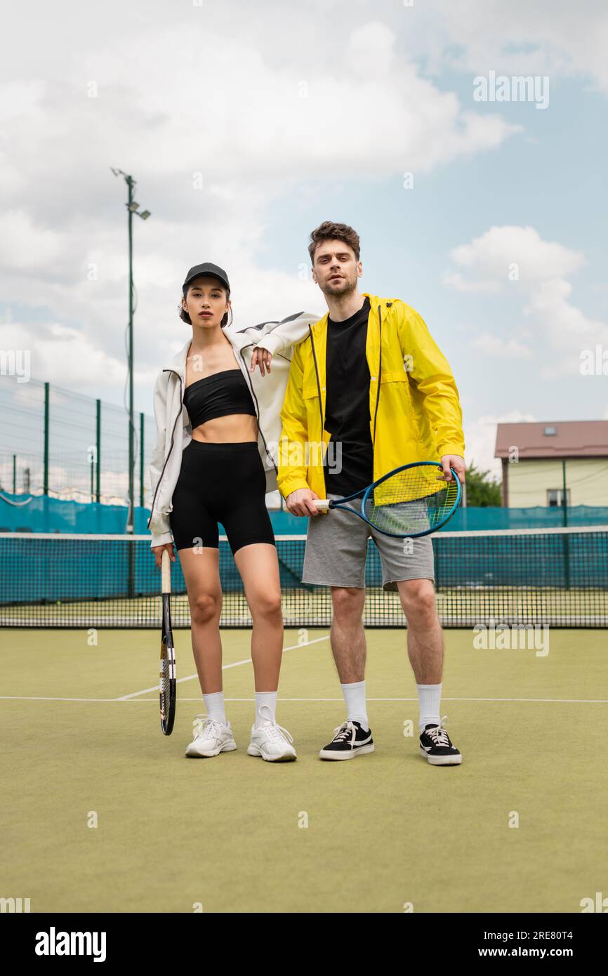 man and woman in stylish active wear standing on court with tennis rackets, healthy lifestyle, sport Stock Photo