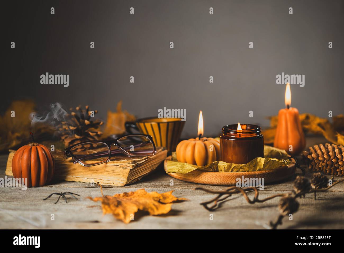 Halloween Still Life on the Wooden Table. Pumpkin Shaped Candles with dry Cones and Leaves, Old Book and Glasses Stock Photo