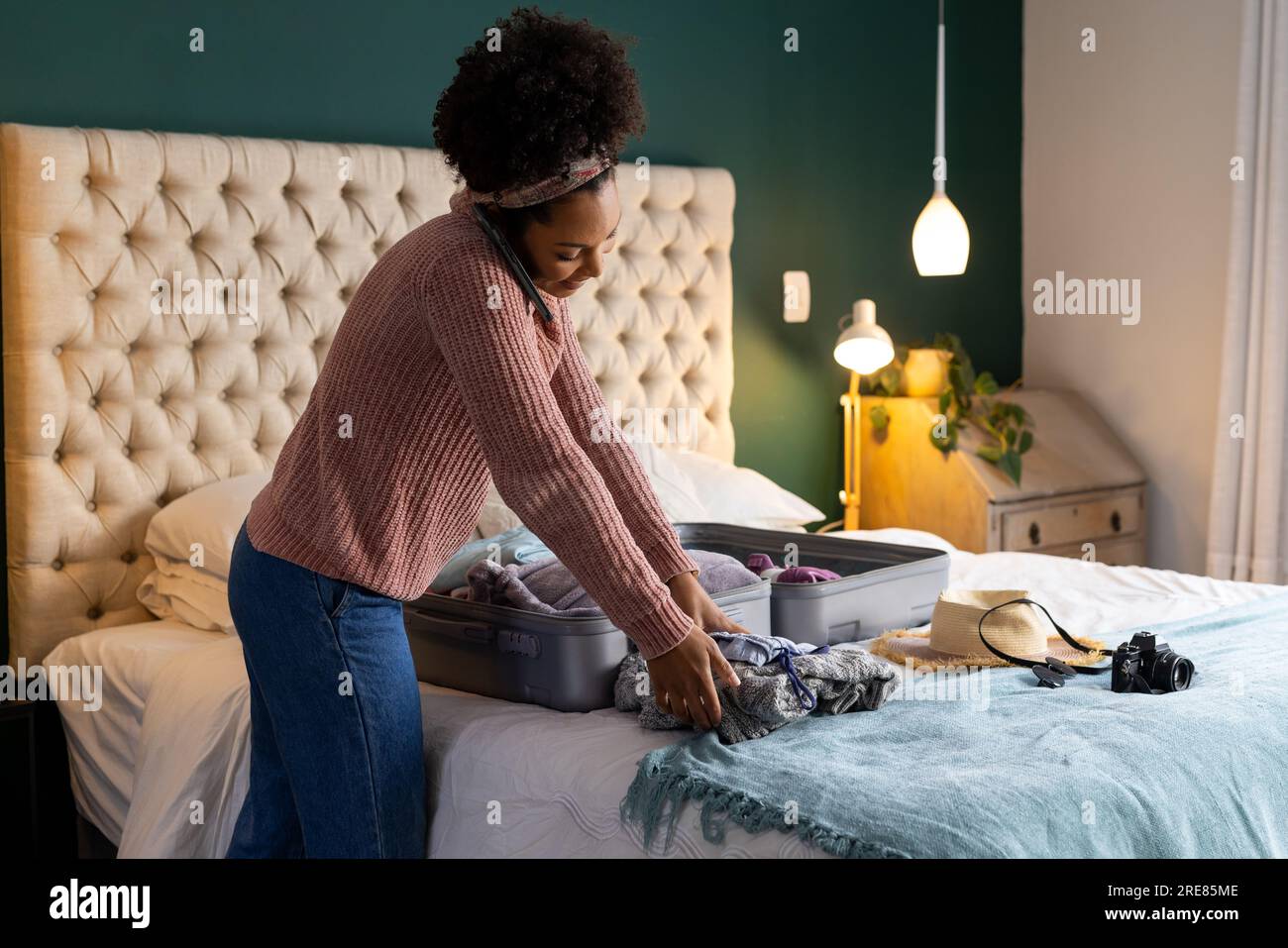 Biracial woman packing suitcase and talking on smartphone in bedroom Stock Photo